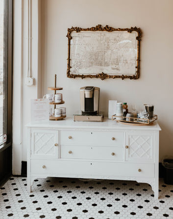 Bright boutique-style coffee station on a white vintage dresser with a single-serve coffee maker, stacked mugs on a tiered stand, tray of stirrers and condiments, ornate framed map on the wall, and black-and-white hex tile floor.