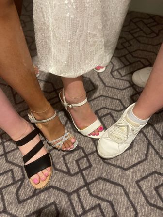 Four women's feet in mixed footwear — black slide sandals, sparkly silver strappy heels, white ankle-strap heels with pink toenails, and a white sneaker — standing on a geometric patterned indoor carpet beneath a sequined dress hem.