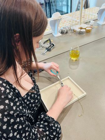 Person assembling a gold chain necklace with pliers at a jewelry-making workstation, surrounded by chain spools, bead trays, display busts and a glass of yellow drink.