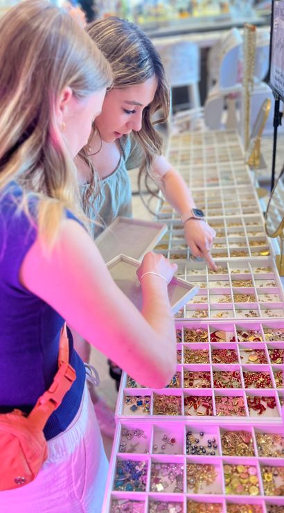 Two friends browsing colorful gold and enamel charms at a boutique jewelry counter, picking pieces from organized display trays under pink-hued store lighting.