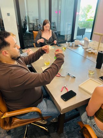 Jewelry-making workshop in a bright studio: people seated around a craft table assembling gold chains and beads with pliers, trays, jewelry displays and drinks nearby.
