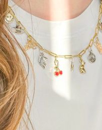 Close-up of a gold paperclip charm necklace with red cherry, fish, bunny, peace sign and assorted charms worn over a white top — trendy layered women's fashion accessory