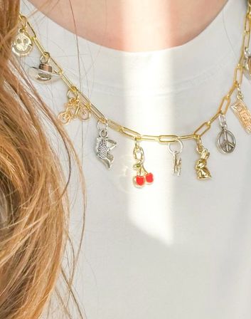 Close-up of a gold paperclip charm necklace with red cherry, fish, bunny, peace sign and assorted charms worn over a white top — trendy layered women's fashion accessory