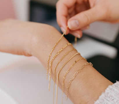 Close-up of a person fastening multiple delicate gold chain bracelets on their wrist — layered bracelet styling, minimalist jewelry photographed in a bright indoor setting.