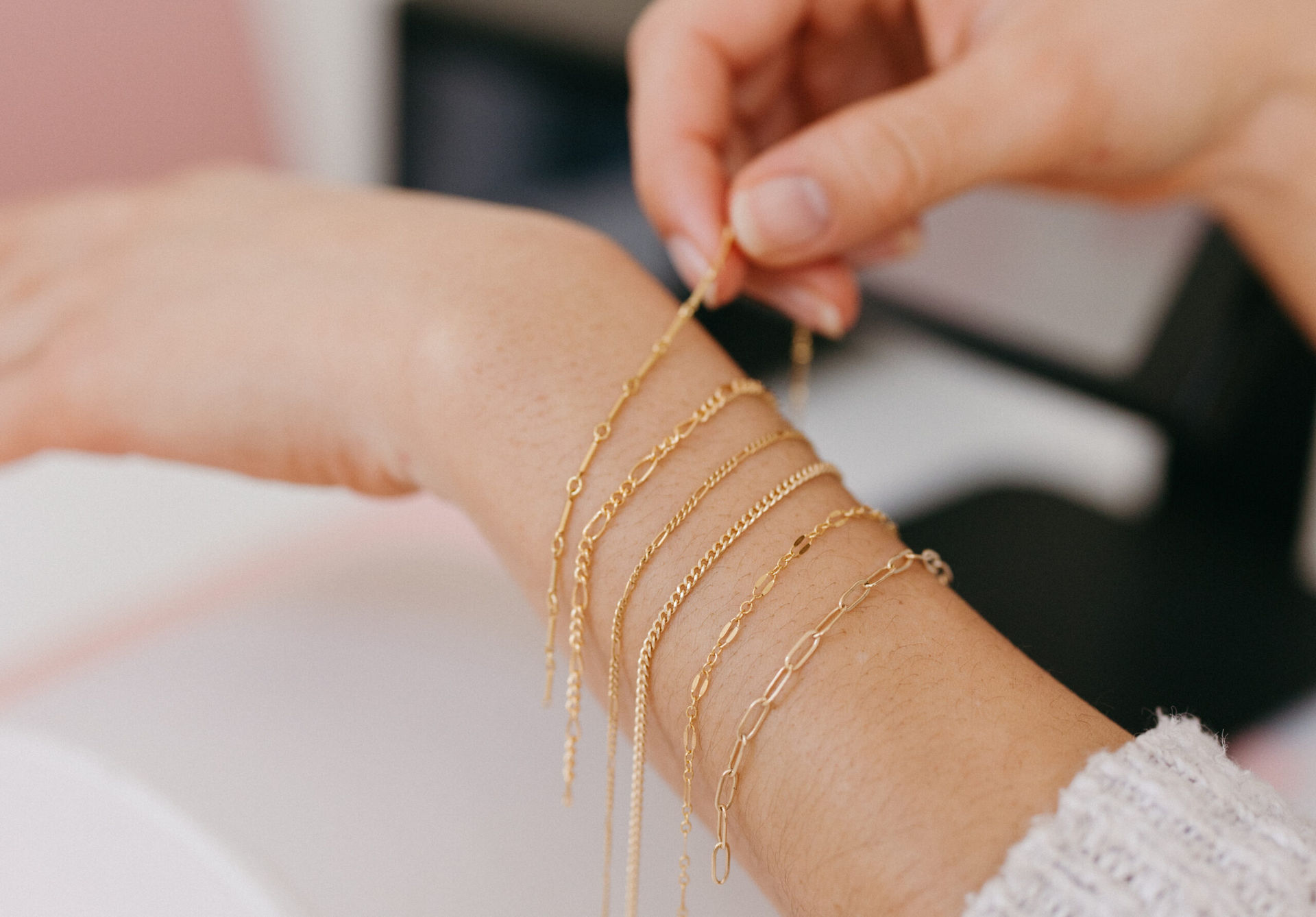 Close-up of a person fastening multiple delicate gold chain bracelets on their wrist — layered bracelet styling, minimalist jewelry photographed in a bright indoor setting.