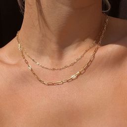 Close-up of tanned neckline and collarbones wearing two layered gold necklaces — a delicate chain and a paperclip-link chain, sunlit minimal jewelry