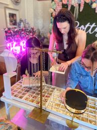 Three women browse a tabletop jewelry display at an indoor boutique pop-up, picking necklaces and rings from white tray organizers and a gold hanging stand beside a round mirror, floral backdrop and pink neon sign.