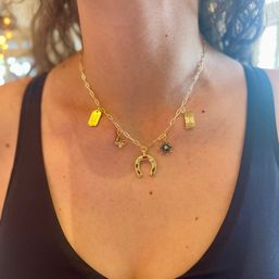 Close-up of a gold charm necklace featuring a horseshoe pendant, star, butterfly and tag charms on a chunky chain, worn at a person's neckline over a black tank top — boho fashion jewelry.