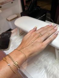 Hand with pink nails and yellow French tips with rhinestone accents, wearing a delicate gold hand‑chain and layered bracelets, resting on a white manicure armrest in a nail salon