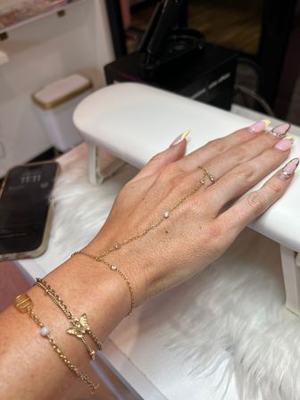 Hand with pink nails and yellow French tips with rhinestone accents, wearing a delicate gold hand‑chain and layered bracelets, resting on a white manicure armrest in a nail salon