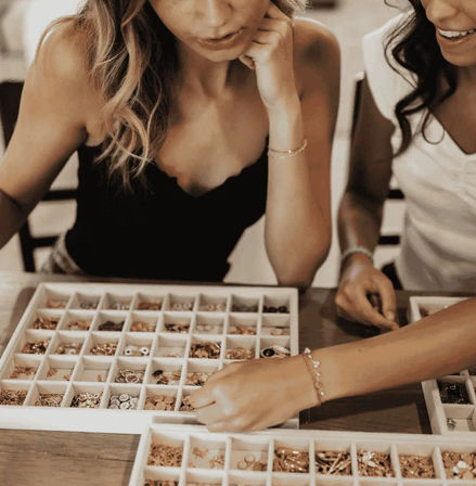 Two women at a wooden table sorting beads, charms and small gold and silver jewelry components from compartment trays during a DIY jewelry-making session.