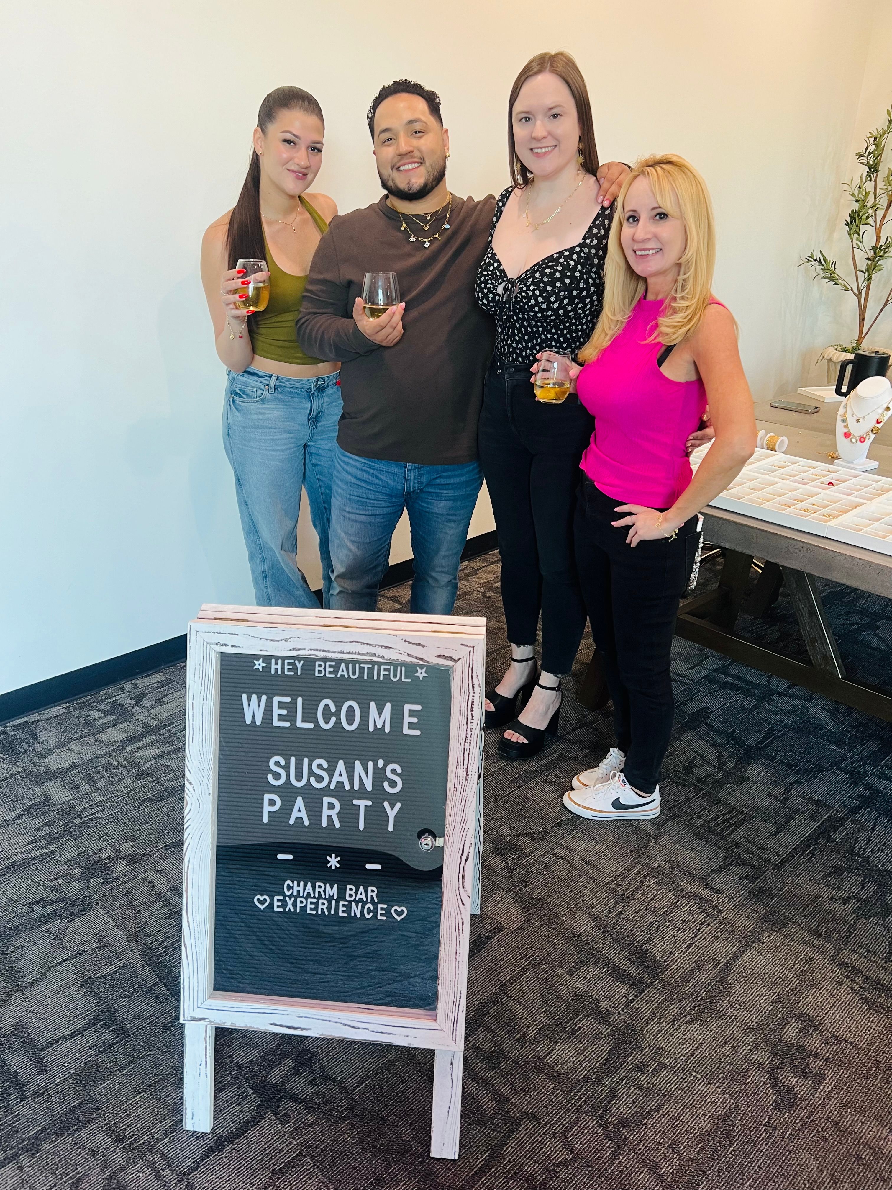Four adults smiling and holding drinks at an indoor jewelry-themed party beside a welcome sign and charm bar display in a bright event space.