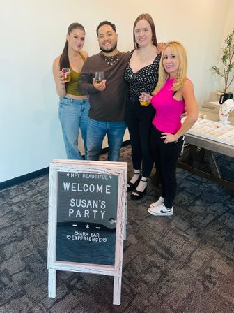 Four adults smiling and holding drinks at an indoor jewelry-themed party beside a welcome sign and charm bar display in a bright event space.