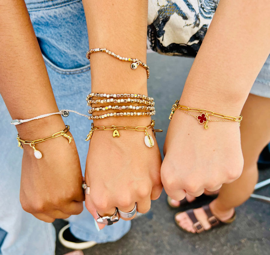 Three wrists displaying stacked gold charm and beaded bracelets with shell and clover charms, paired with casual denim and sandals — boho summer street-style friendship jewelry.