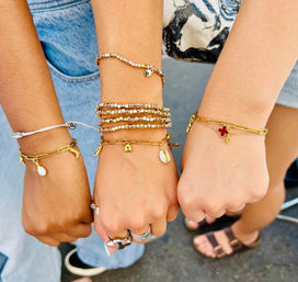 Three wrists displaying stacked gold charm and beaded bracelets with shell and clover charms, paired with casual denim and sandals — boho summer street-style friendship jewelry.