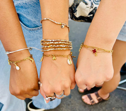 Three wrists displaying stacked gold charm and beaded bracelets with shell and clover charms, paired with casual denim and sandals — boho summer street-style friendship jewelry.