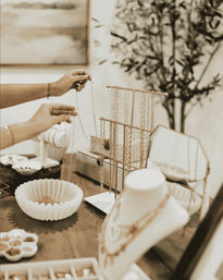 Hands arranging gold necklaces on a tabletop boutique jewelry display with a chain rack, display bust, mirror and small bowls of accessories