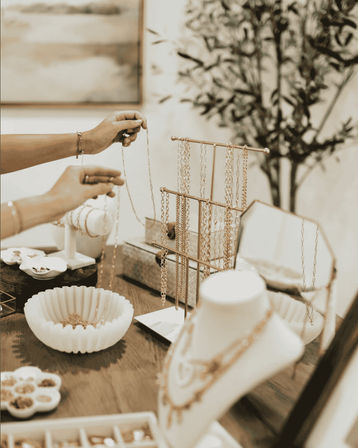Hands arranging gold necklaces on a tabletop boutique jewelry display with a chain rack, display bust, mirror and small bowls of accessories