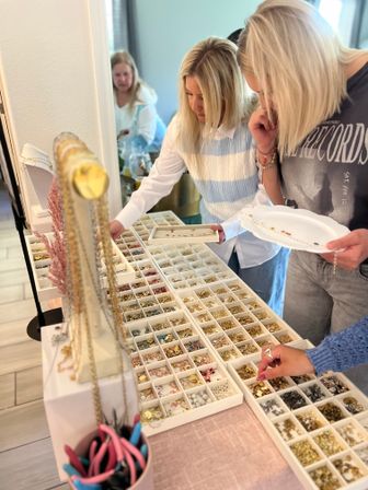 Three women at an indoor DIY jewelry station sorting beads, charms, and findings from organized white trays with necklaces on display and pliers nearby
