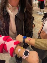 Close-up in a clothing boutique: three wrists showing smartwatches and gold heart charm bracelets, layered charm necklace, pink polka-dot sleeve and black leather jacket, wooden floor and clothing racks in background.
