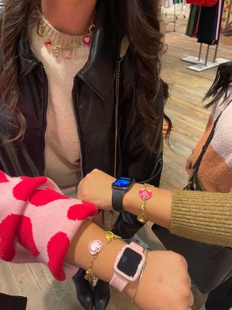 Close-up in a clothing boutique: three wrists showing smartwatches and gold heart charm bracelets, layered charm necklace, pink polka-dot sleeve and black leather jacket, wooden floor and clothing racks in background.