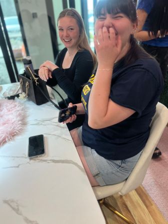 Two women laughing at a marble-topped table in a bright modern cafe, one holding a smartphone with a black handbag nearby