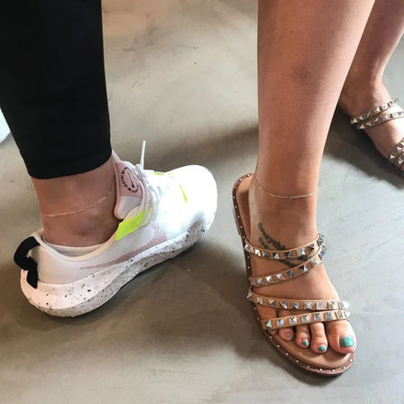 Two ankles with delicate chain anklets—one in a white neon-accented sneaker, the other in a studded strappy sandal showing teal toenails and a small foot tattoo, standing on a smooth concrete floor.