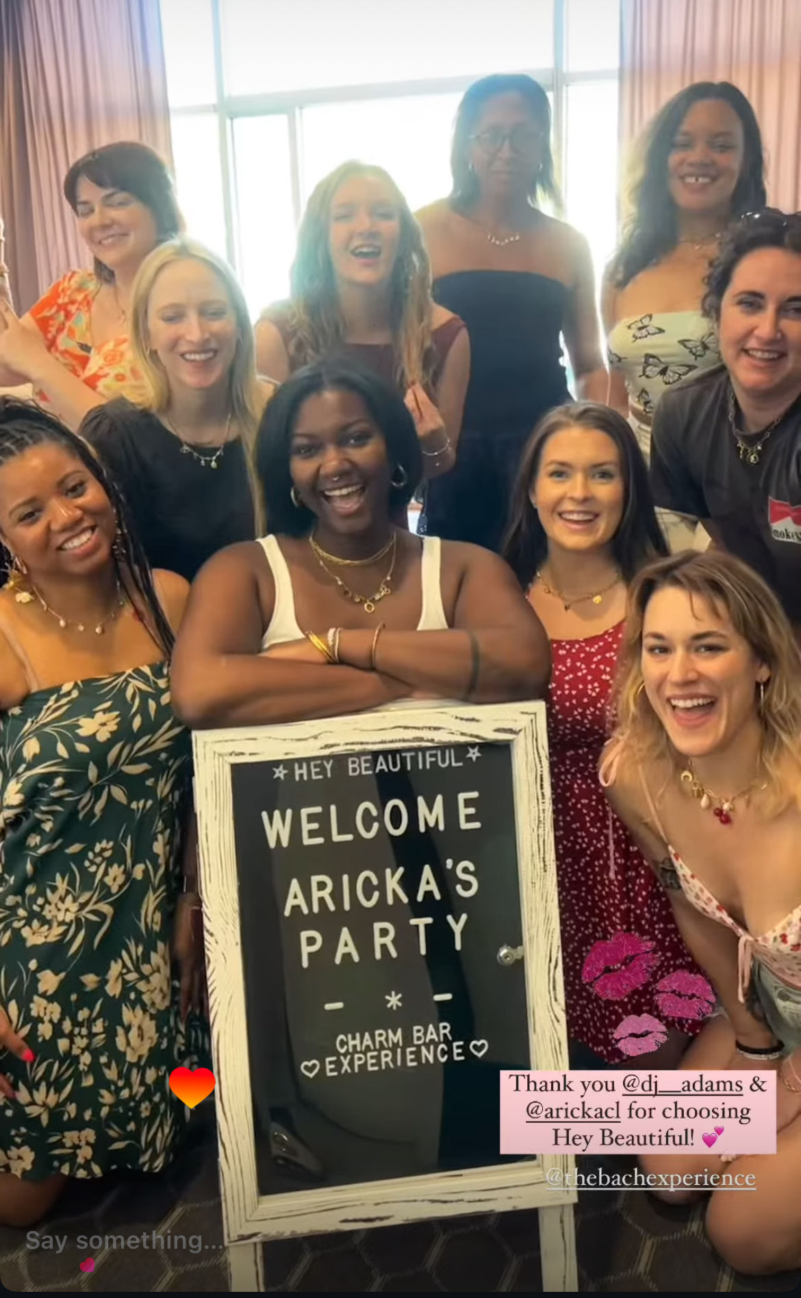 Smiling diverse group of women gathered in a sunlit room, posing around a rustic framed welcome sign at a festive party, wearing summer dresses and jewelry.