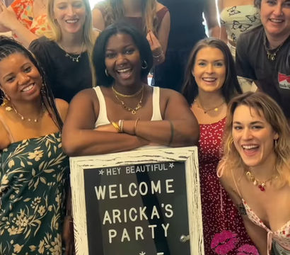 Smiling diverse group of women gathered in a sunlit room, posing around a rustic framed welcome sign at a festive party, wearing summer dresses and jewelry.