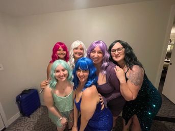 Six friends wearing bright wigs (pink, white, mint, blue, lavender, black) and sparkly dresses smiling and posing for a group photo in an indoor hotel room setting