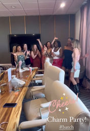 Group of women cheering and clapping in a hotel meeting room, gathered near a wooden conference table with beige chairs and a wall-mounted TV, holding a bottle of champagne — indoor celebration/party.