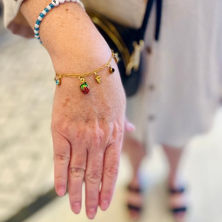 Close-up of a hand wearing a gold charm bracelet with a red strawberry charm and a blue beaded bracelet against a blurred indoor background