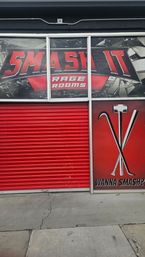 Red storefront exterior with closed roll-up door, shattered-glass rage-room graphics and crossed sledgehammer, crowbar and bat icons above an urban sidewalk.