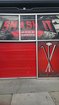 Red storefront exterior with closed roll-up door, shattered-glass rage-room graphics and crossed sledgehammer, crowbar and bat icons above an urban sidewalk.