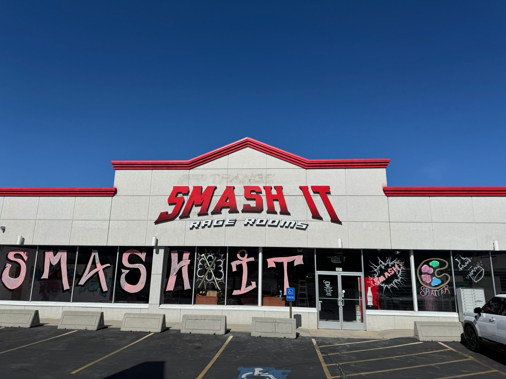 Strip-mall storefront for a smash-themed rage room with bold red 3D signage, painted window letters and splatter artwork, empty parking spaces under a clear blue sky.