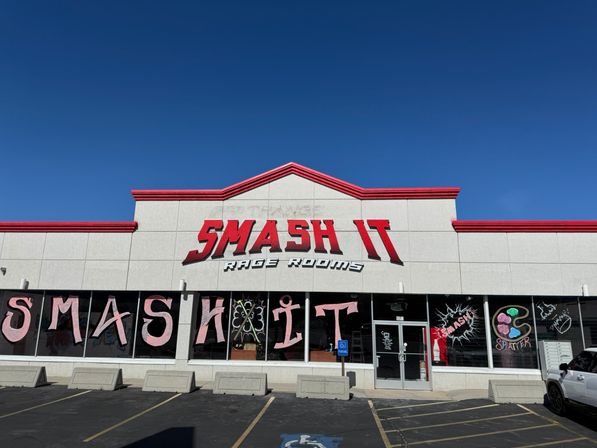 Strip-mall storefront for a smash-themed rage room with bold red 3D signage, painted window letters and splatter artwork, empty parking spaces under a clear blue sky.