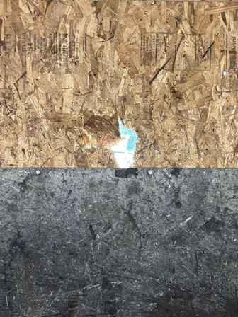Boarded construction surface: rough OSB plywood with a small tear revealing bright blue behind, above a dark weathered asphalt/tar panel with visible screws.