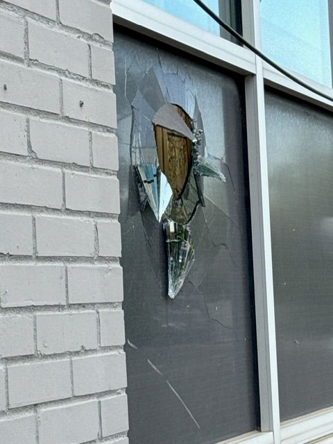 Shattered storefront window with jagged broken glass and hanging shards in a gray-tinted pane beside a white brick exterior