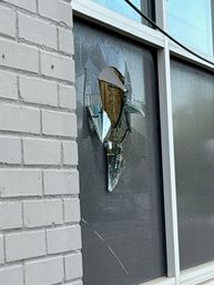 Shattered storefront window with jagged broken glass and hanging shards in a gray-tinted pane beside a white brick exterior
