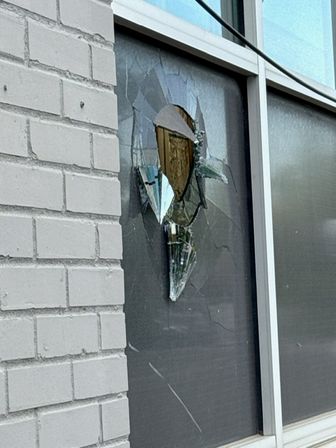Shattered storefront window with jagged broken glass and hanging shards in a gray-tinted pane beside a white brick exterior