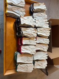 Bulk stacks of white leather work gloves with colored cuffs (green, red, yellow, black) neatly arranged on a wooden table next to cardboard boxes in a workshop