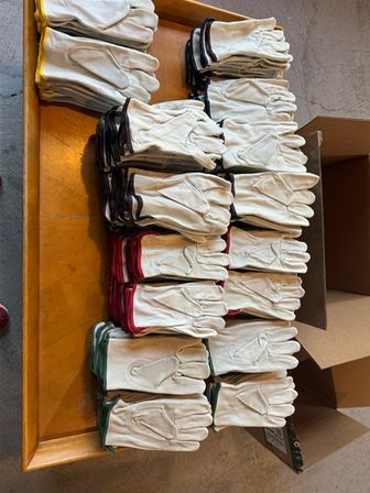 Bulk stacks of white leather work gloves with colored cuffs (green, red, yellow, black) neatly arranged on a wooden table next to cardboard boxes in a workshop