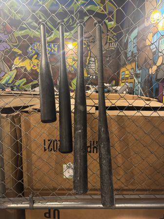 Four black baseball bats hanging on a chain-link fence in front of stacked cardboard boxes, colorful graffiti mural and industrial lighting in an indoor urban sports space.