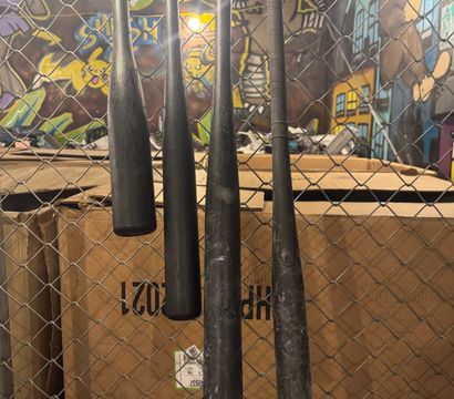 Four black baseball bats hanging on a chain-link fence in front of stacked cardboard boxes, colorful graffiti mural and industrial lighting in an indoor urban sports space.
