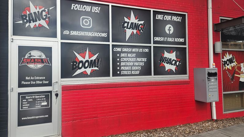 Red brick storefront with comic-book style rage-room window graphics showing words BANG, BOOM, CLANG, WHACK and POW; window sign lists date night, corporate parties, birthday parties, private events and stress-relief sessions.