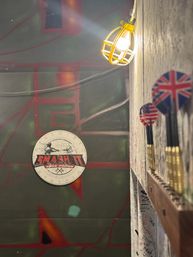 Pub-style indoor darts area with circular dartboard on a graffiti wall, row of darts with UK and US flag flights in a wooden rack, and a glowing yellow caged wall light