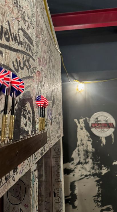 Close-up of darts with Union Jack and U.S. flag flights on a wooden rack against a graffiti-covered wall in an indoor bar game area with an exposed hanging bulb.