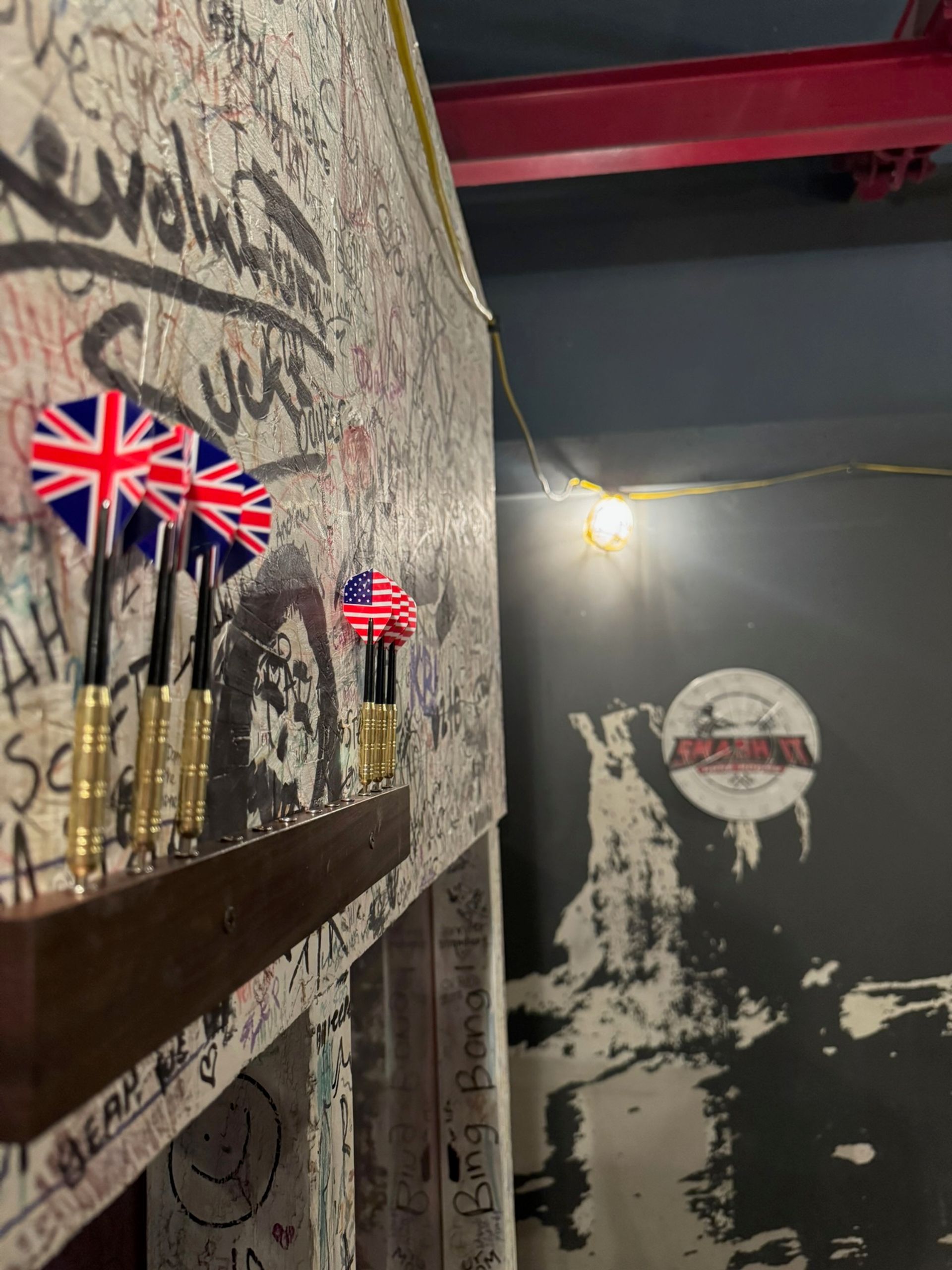 Close-up of darts with Union Jack and U.S. flag flights on a wooden rack against a graffiti-covered wall in an indoor bar game area with an exposed hanging bulb.