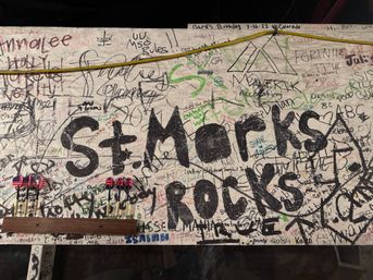 Graffiti-covered wooden pub wall packed with handwritten signatures, doodles and a large black-painted slogan, two sets of darts with American and British flag flights on a wooden rack, and a yellow cable strung above.