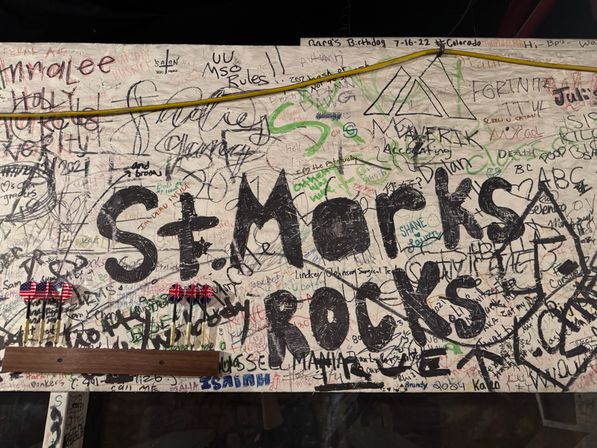 Graffiti-covered wooden pub wall packed with handwritten signatures, doodles and a large black-painted slogan, two sets of darts with American and British flag flights on a wooden rack, and a yellow cable strung above.